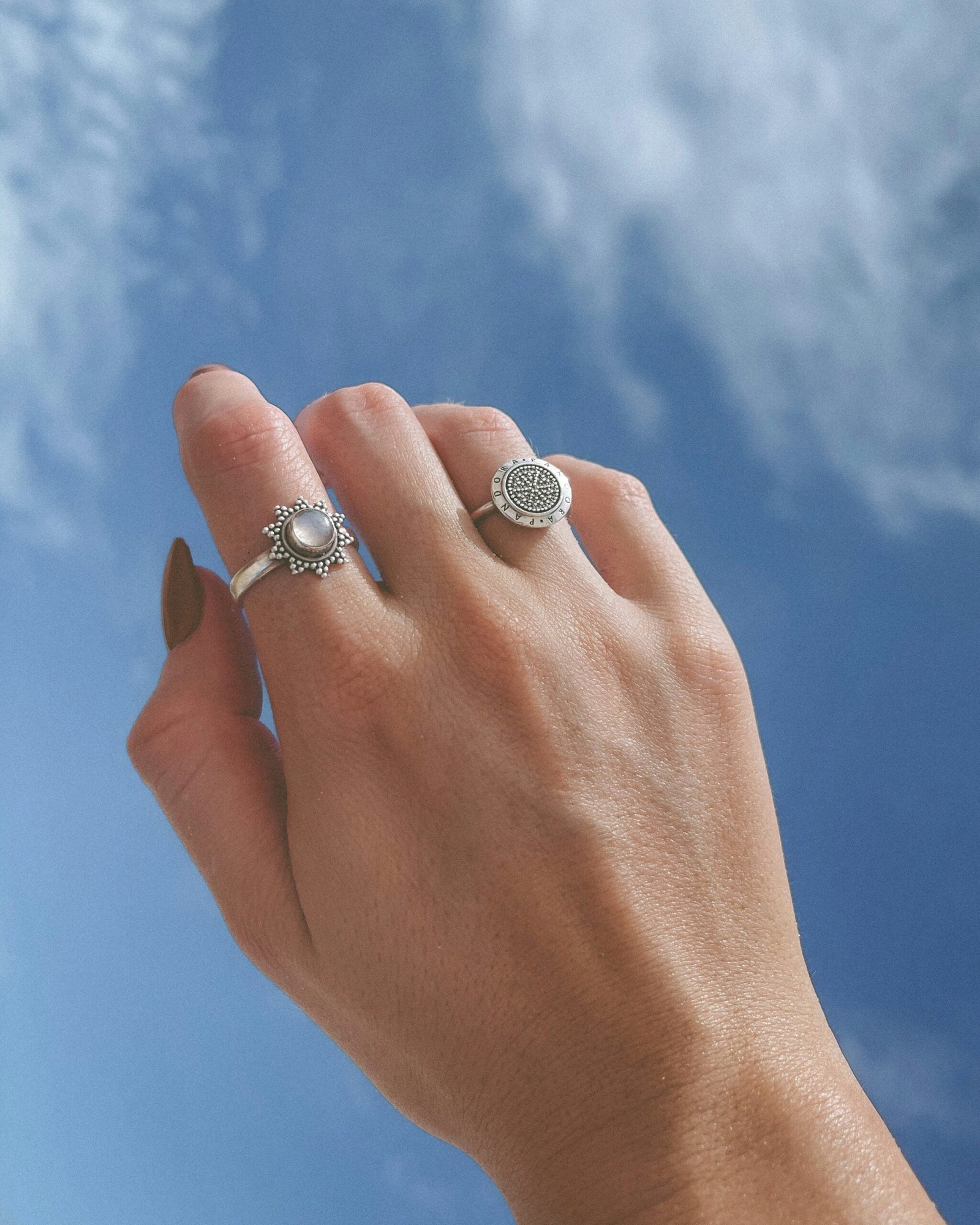 Muestra una mano alzada con un fondo de cielo azul con algunas nubes dispersas, se pueden ver dos anillos de plata en la mano:
el anillo en el dedo medio tiene un diseño ornamental con un gran elemento central circular, que parece una piedra o adorno brillante rodeado por pequeños detalles decorativos. Tiene un estilo elegante y vintage. Anillo de plata en el dedo anular: tiene una forma circular en el centro, pero su diseño es más sencillo y moderno en comparación con el anterior. Alrededor del círculo central se observa un patrón detallado, que podría ser textura o pequeños grabados.