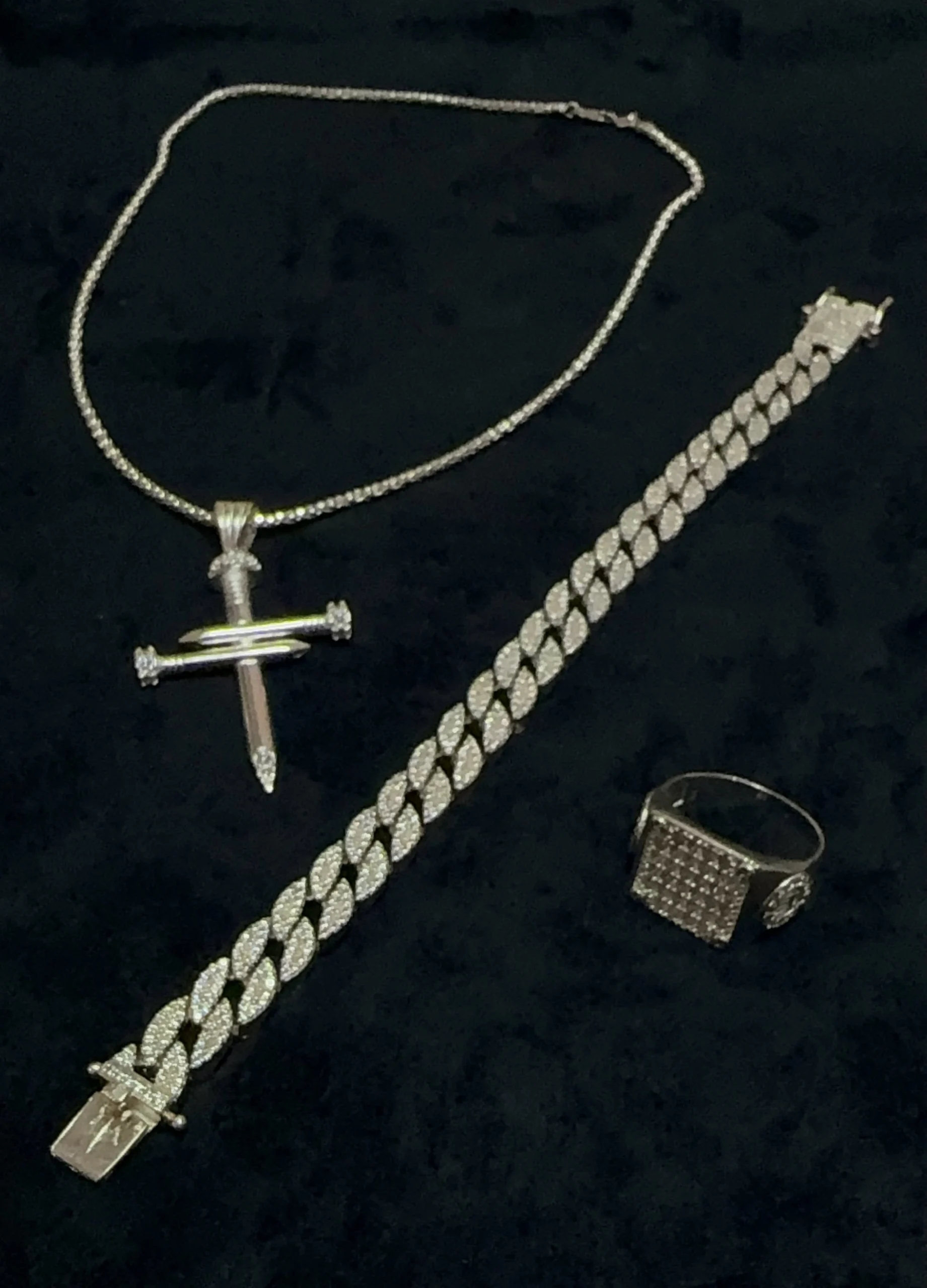 Joyería para hombre en plata con incrustaciones de diamantes, collar con cruz, pulsera y anillo sobre terciopelo negro.