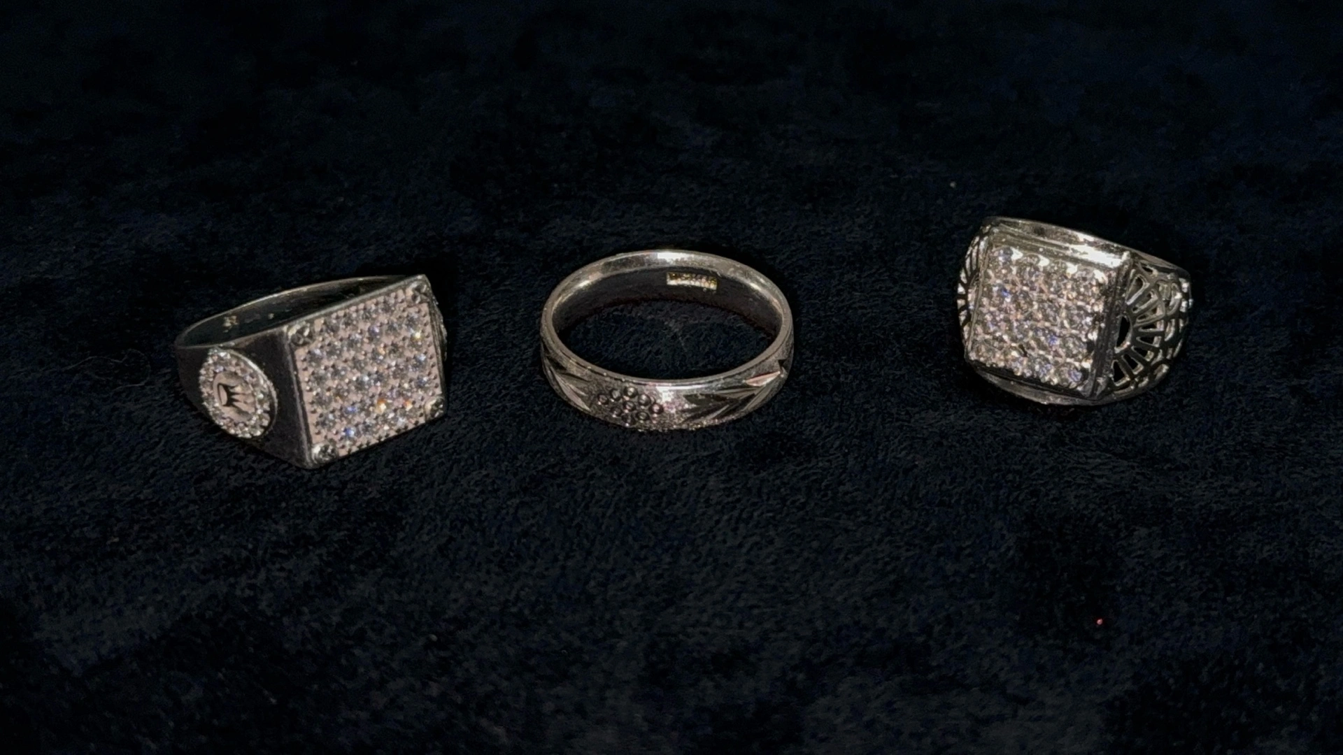 Colección de anillos algunos con incrustaciones de pequeñas piedras brillantes como zafiros azules o rubies rojos, anillos de plata con forma de corazón o flor con alguna piedra central o rodeada con algunas de menor tamaño, y muchos otros sin piedras solo con acabados lindos y elegantes, también se pueden ver brazaletes con diseño moderno al que puedes agregar colgantes de joyería para hacerlos individual y distintivos. Todo esto se encuentra en una estructura en filas sobre una superficie gris aterciopelada.