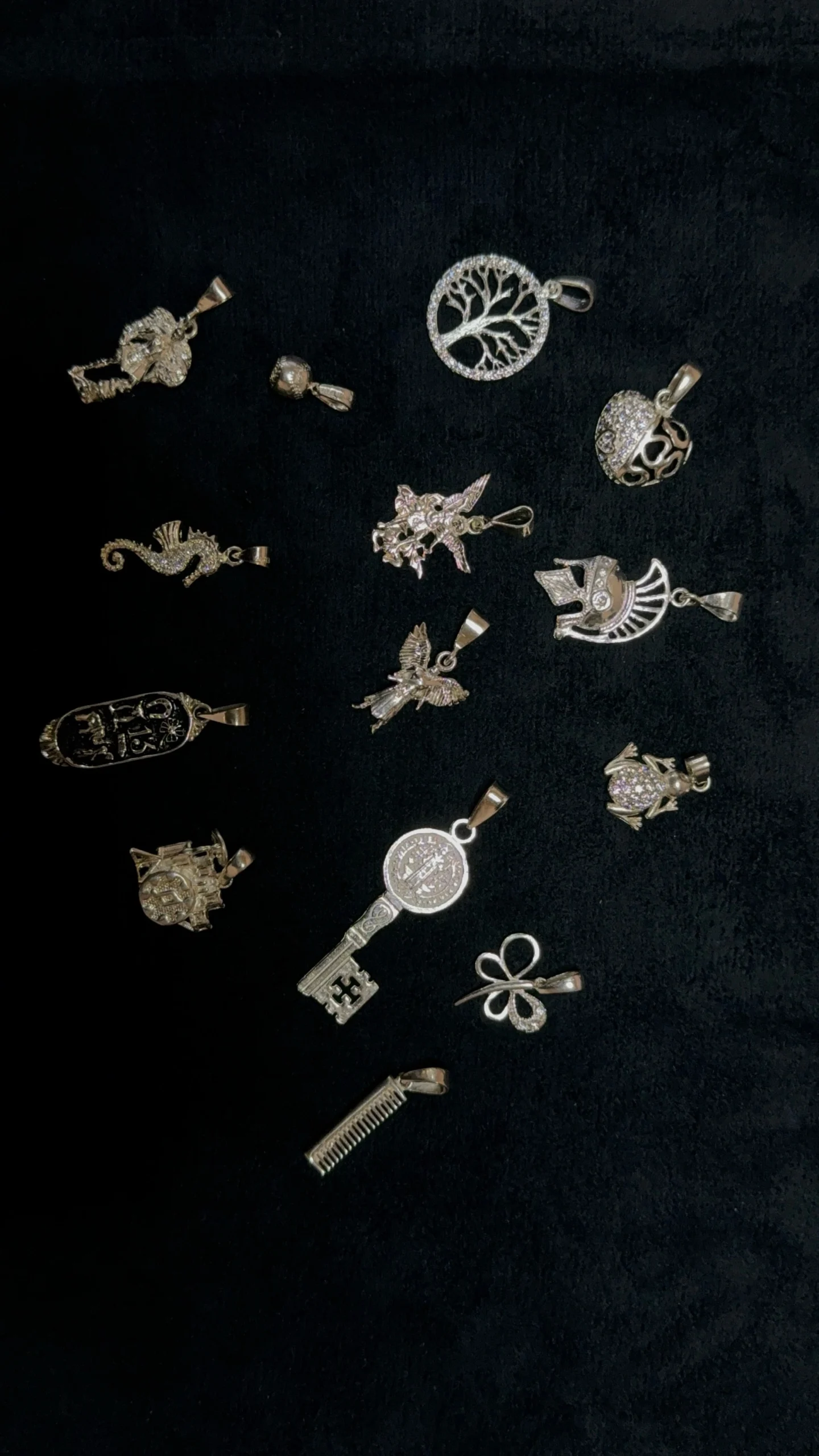 Colección de dijes de plata con diferentes diseños, incluyendo un árbol de la vida, una llave, una mariposa y varios animales, sobre un fondo de terciopelo negro. Destaca la variedad y originalidad de los diseños, ideales para personas que buscan dijes únicos y especiales.