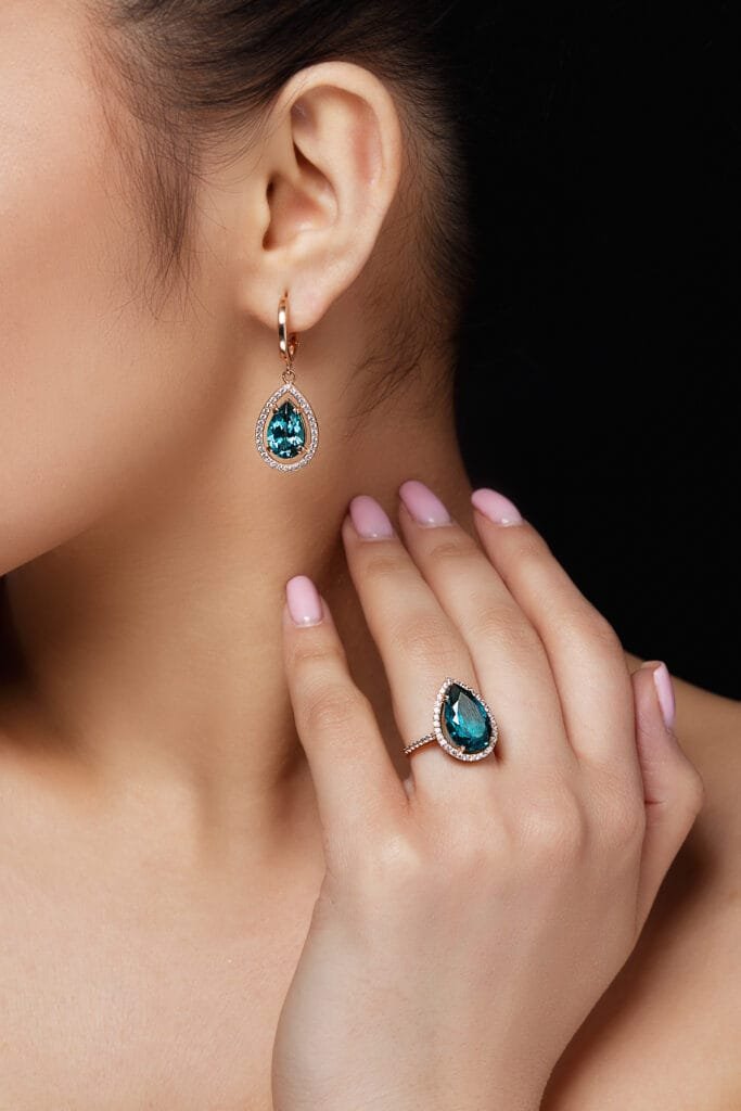Mujer joven usando aretes y anillo de plata con incrustaciones de piedras preciosas en forma de gota con un tono azul verdoso en conjunto de joyería fina, versátil, elegantes y de calidad ideales para mujeres de estilo refinado y delicado.